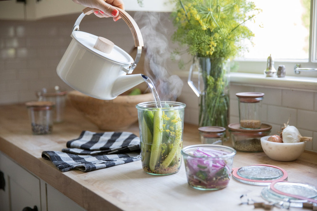 Hands pouring steaming water from a white kettle into a jar of cucumbers and dill for quick pickles on a sunlit kitchen counter