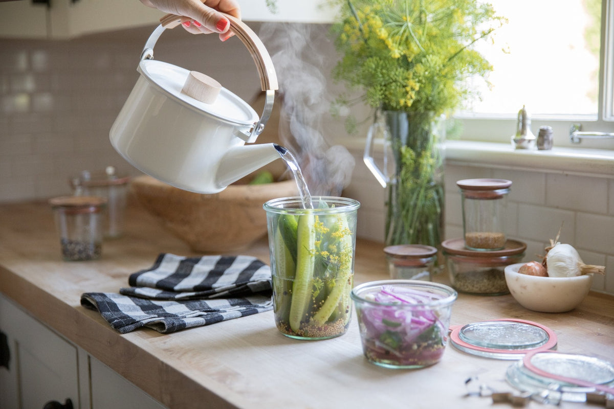 Hands pouring steaming water from a white kettle into a jar of cucumbers and dill for quick pickles on a sunlit kitchen counter