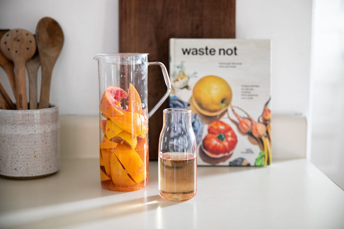 Pitcher of citrus peels and a small carafe of infused vinegar on a kitchen counter with the ‘waste not’ cookbook and wooden utensils in the background