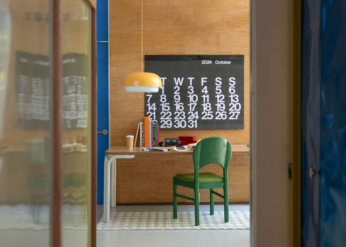 Green chair at a desk in a study nook with a black October 2024 wall calendar, yellow pendant lamp, stacked books, and a coffee cup