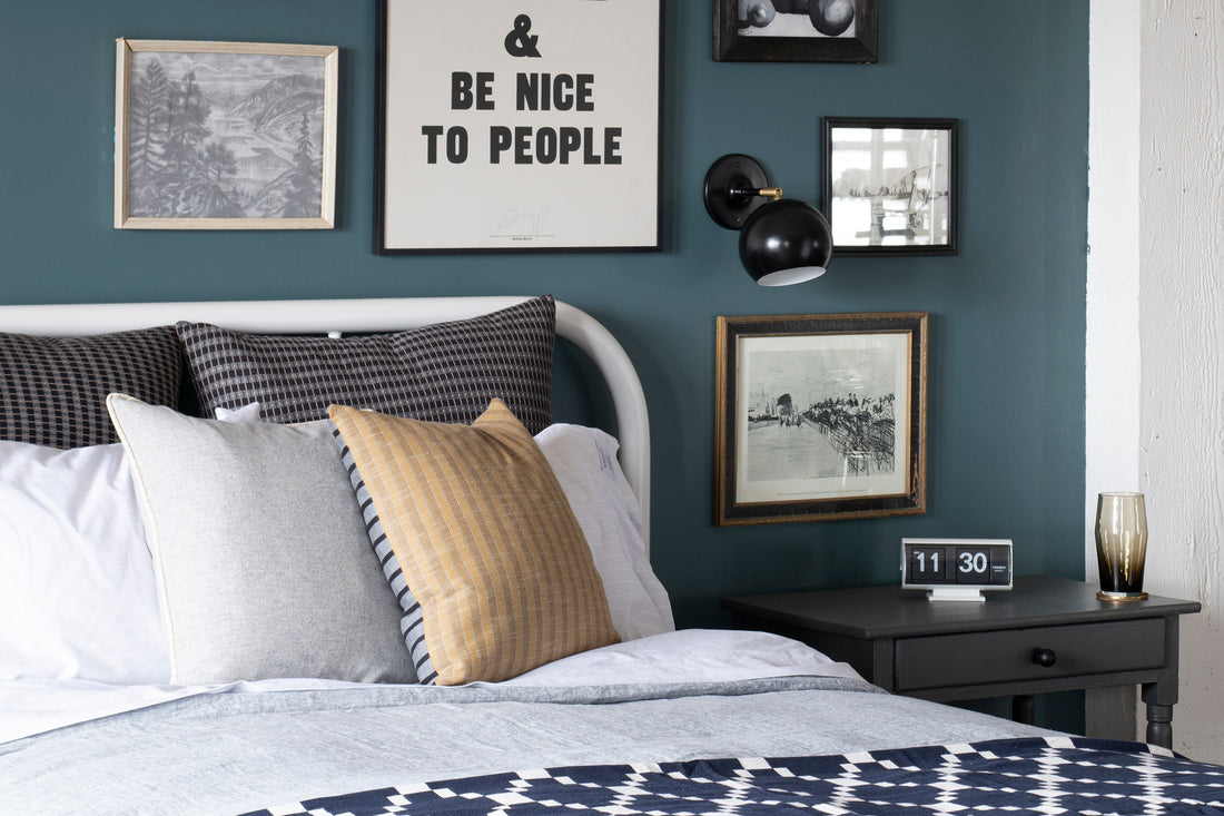 Gallery wall above bed with framed print reading ‘BE NICE TO PEOPLE’, black wall sconce, patterned pillows, and bedside table with flip clock
