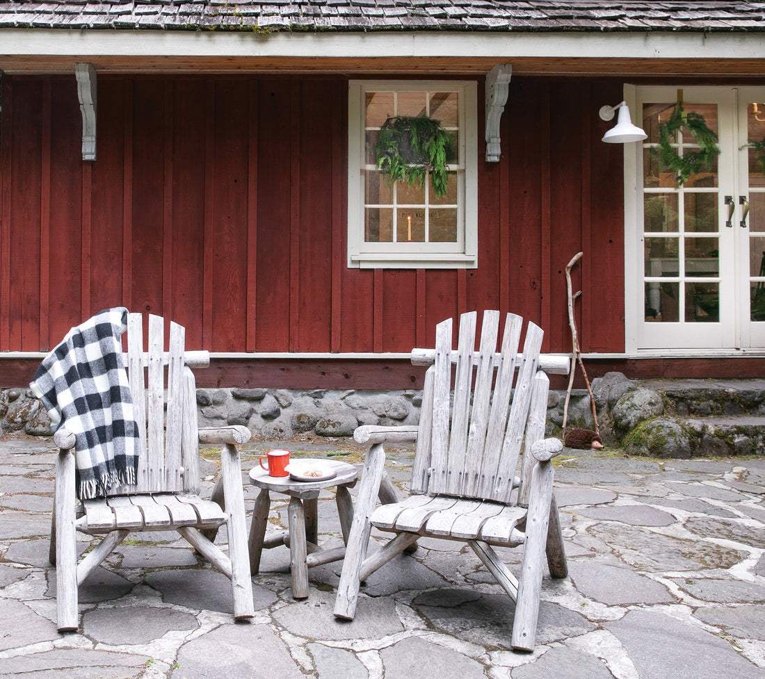 Two weathered wooden chairs and a small table with a red mug and pastry on a stone patio outside a red cottage