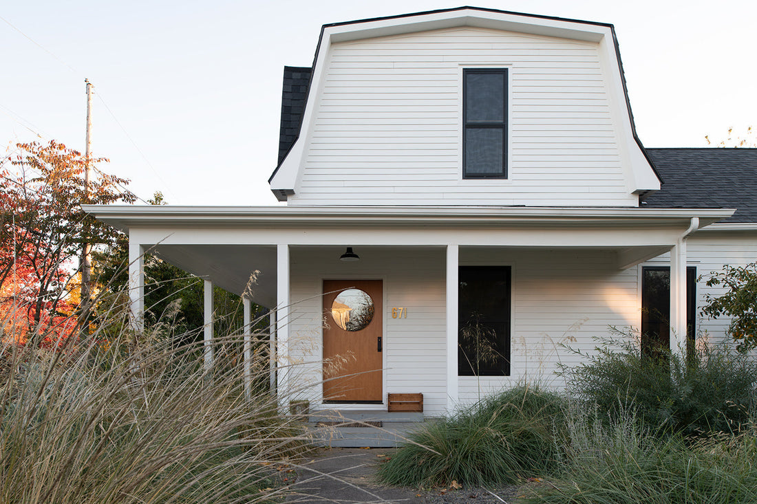 White two-story house with covered front porch, round-window wooden door and ornamental grasses in the yard