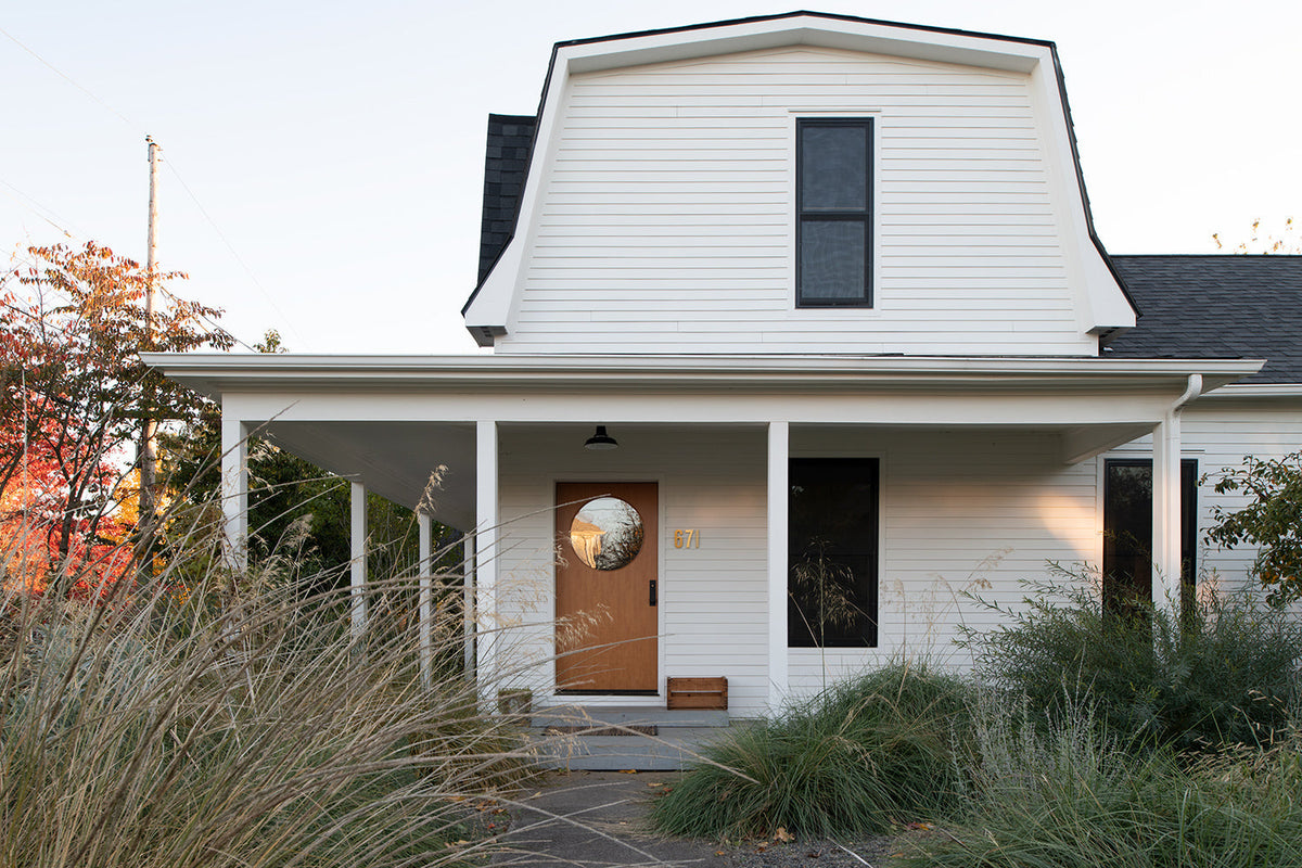 White two-story house with covered front porch, round-window wooden door and ornamental grasses in the yard