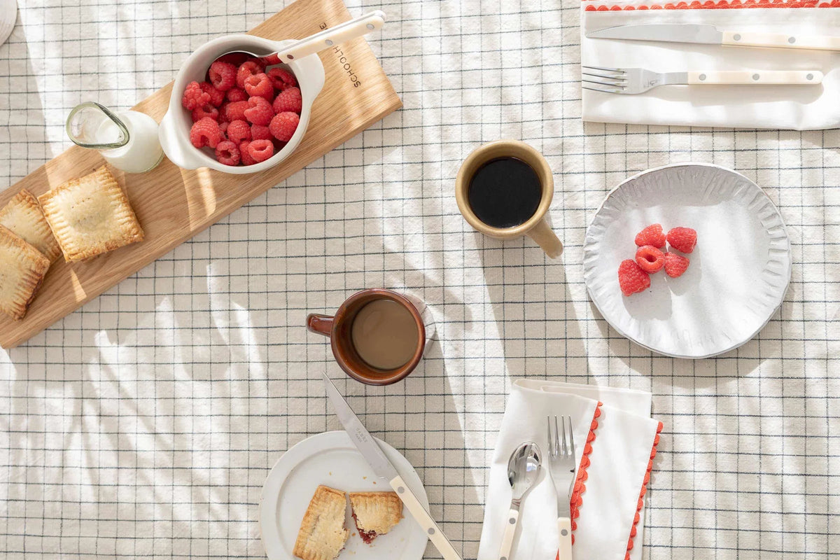 Overhead summer brunch table with raspberries, hand pies on a wood board, two coffee mugs, and napkins with red trim on a checked cloth