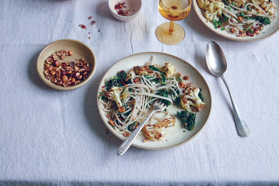 Overhead view of roasted cauliflower, Swiss chard and hazelnut pasta on a ceramic plate with a fork, a bowl of chopped hazelnuts, and a silver serving spoon
