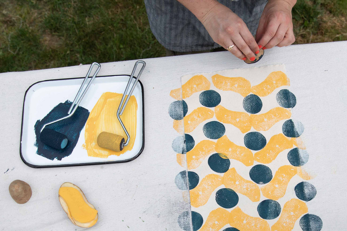 Hands stamping mustard and navy potato block prints on a cream canvas at an outdoor table
