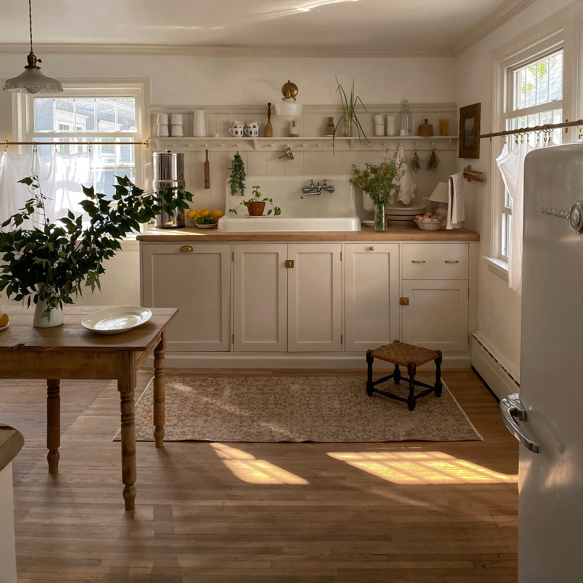 Sunlit vintage kitchen with farmhouse sink, cream cabinetry, wood countertops, potted plants, open shelving, and a small woven stool