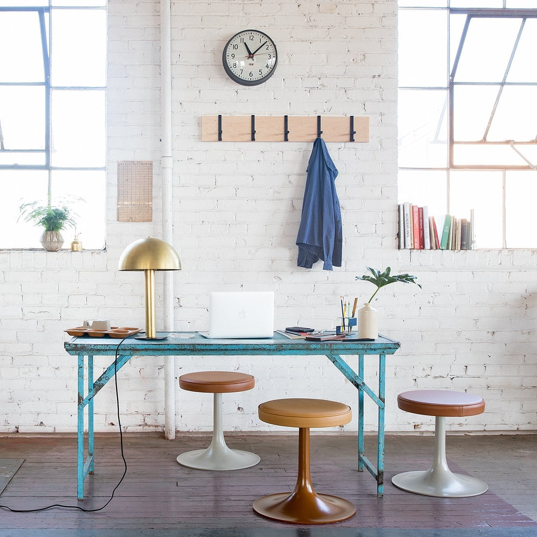 Sunlit studio workspace with turquoise table, brass lamp, three tulip stools, laptop, and coat on wall hooks