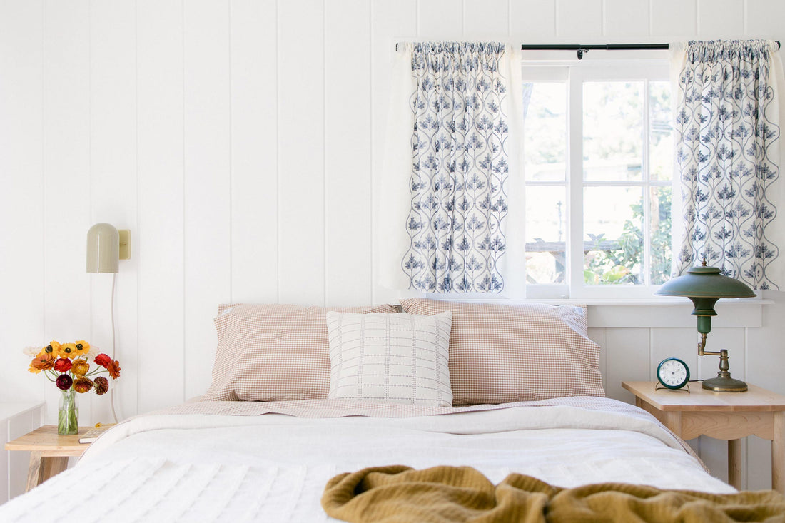 Bedside with plug-in brass wall sconce and green table lamp on wooden nightstands by a window with blue patterned curtains