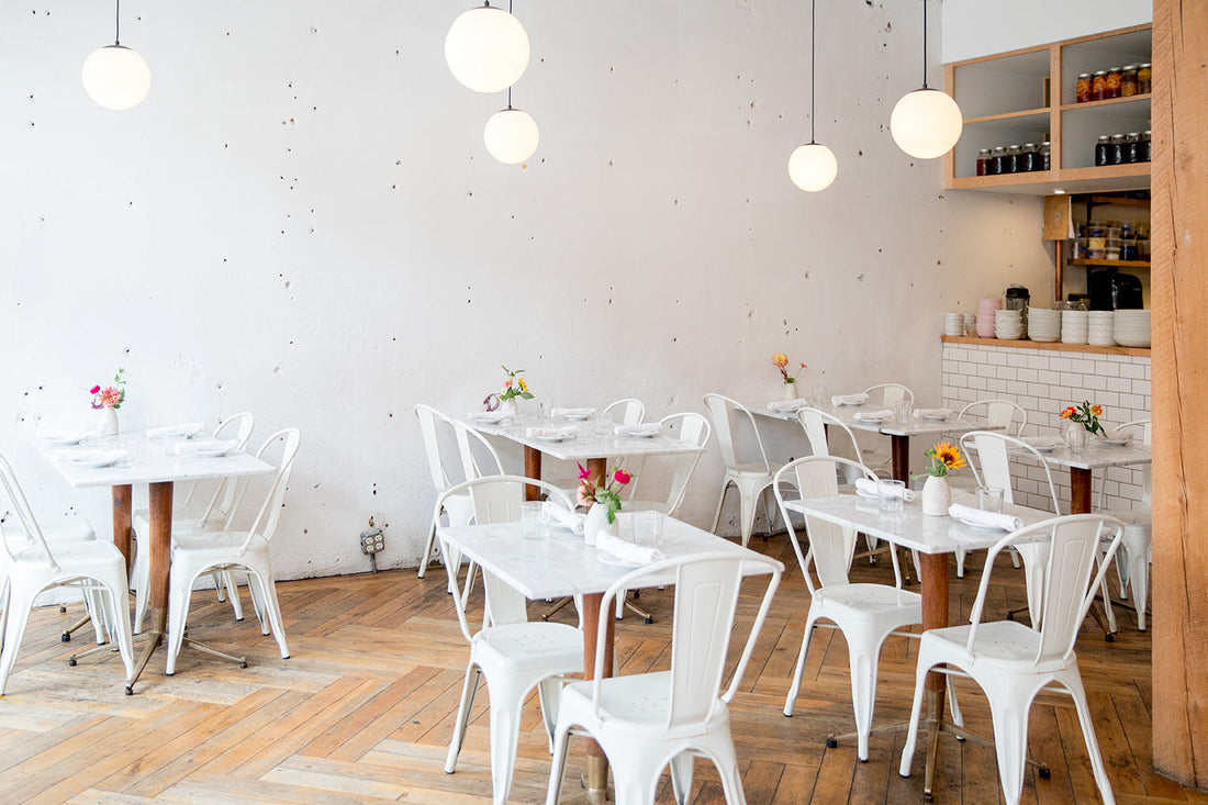 Empty sunlit café interior with white marble tables, white metal chairs, globe pendant lights, herringbone wood floor, and small vases of flowers