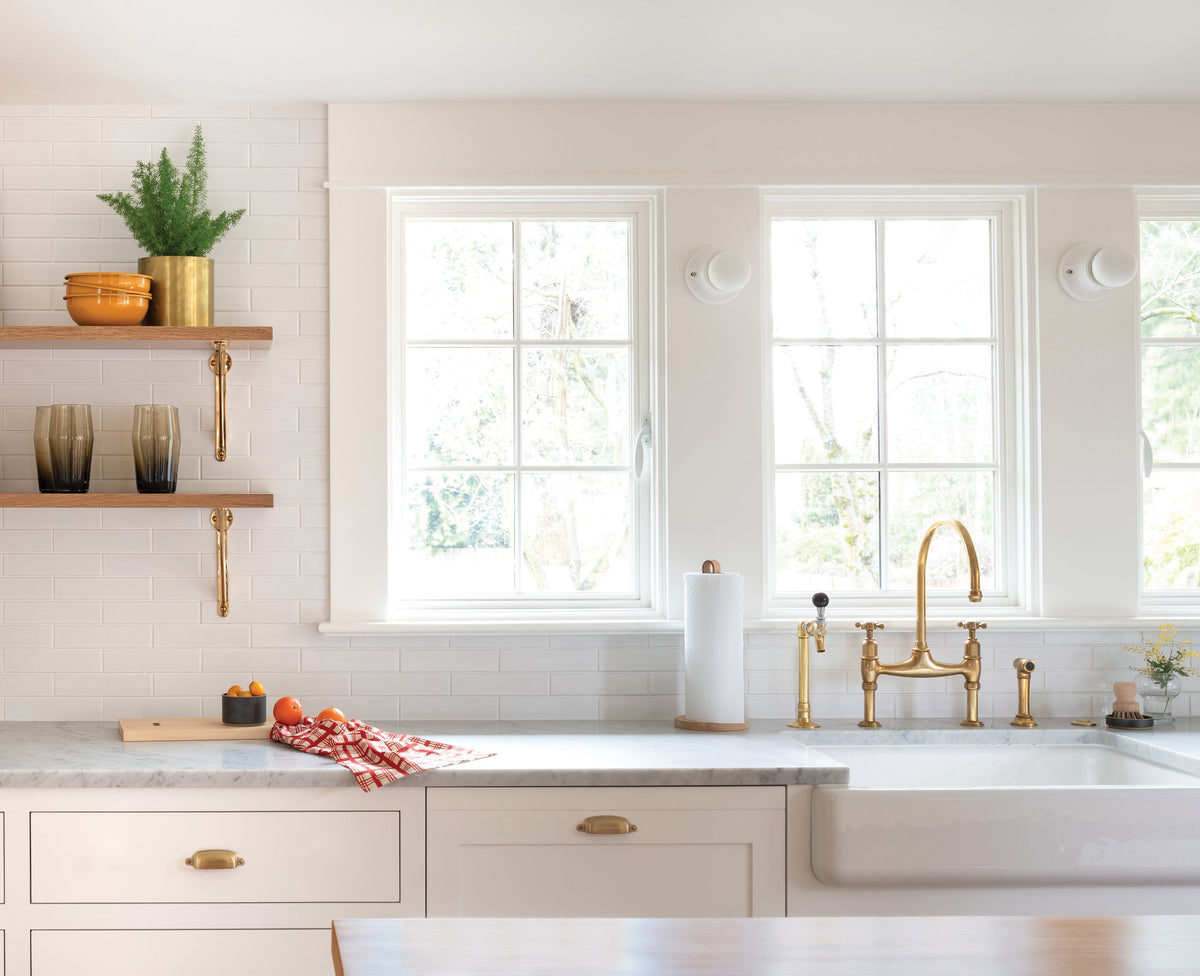 Sunlit farmhouse kitchen sink area with marble countertop, brass bridge faucet, wood floating shelves and a bowl of oranges