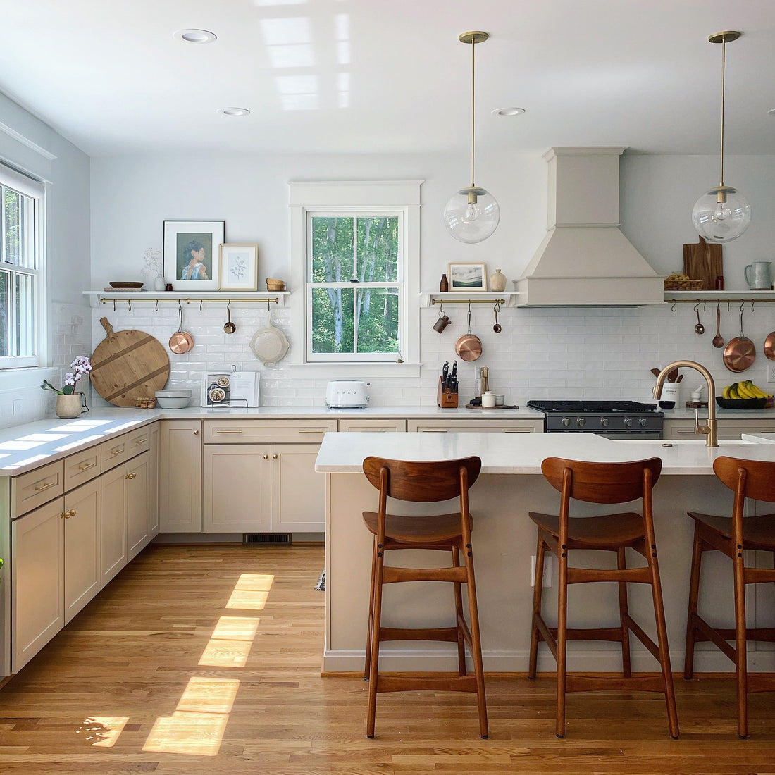 Sunlit farmhouse kitchen with marble island and three wooden bar stools, white cabinets, brass pendants and hanging copper pans