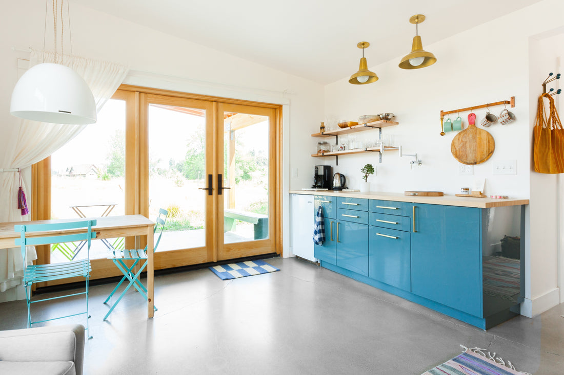 Sunlit farmhouse kitchen with blue lower cabinets, wooden countertop, open glass doors to a backyard, and turquoise bistro chairs