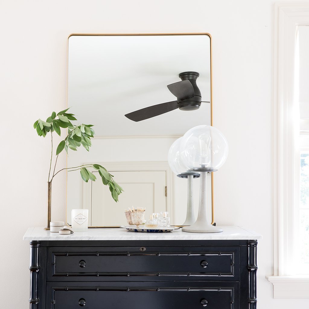 Front view of Leo mirror showcasing thin brass frame and rounded corners with ceiling fan reflection above a black marble-top dresser