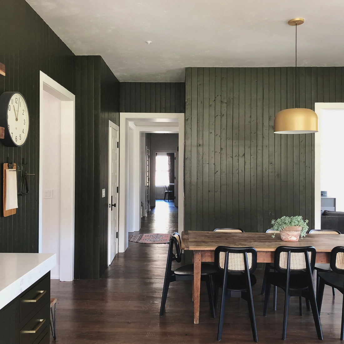 Dining area with long wooden table, black cane-back chairs, dark vertical wood-paneled wall and brass pendant light, hallway view to sunlit room