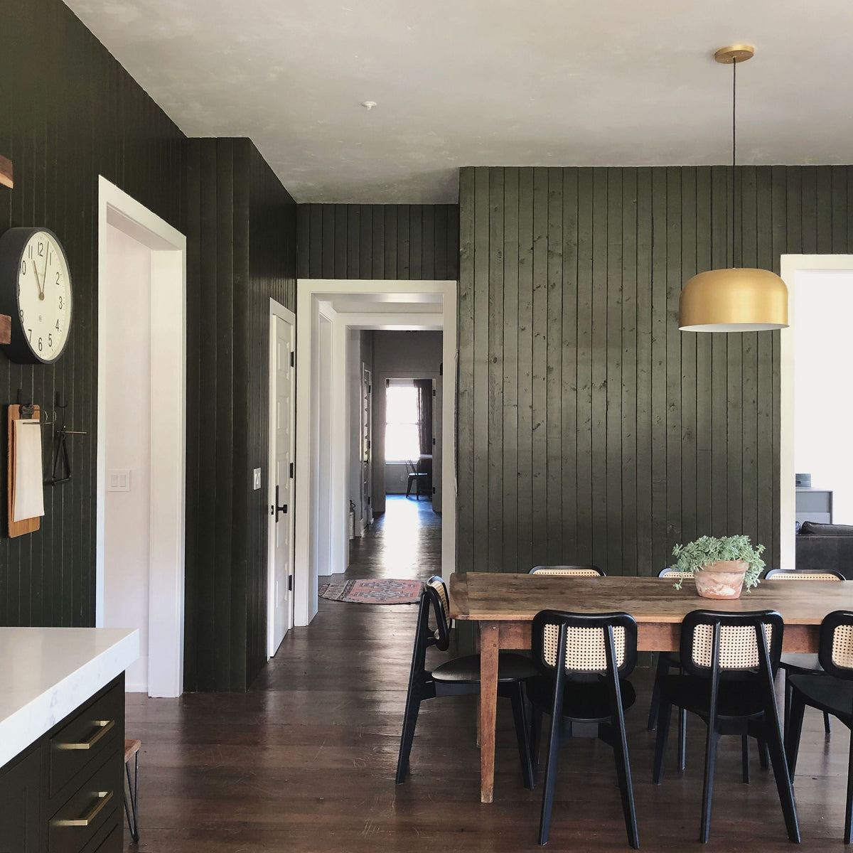 Dining area with long wooden table, black cane-back chairs, dark vertical wood-paneled wall and brass pendant light, hallway view to sunlit room