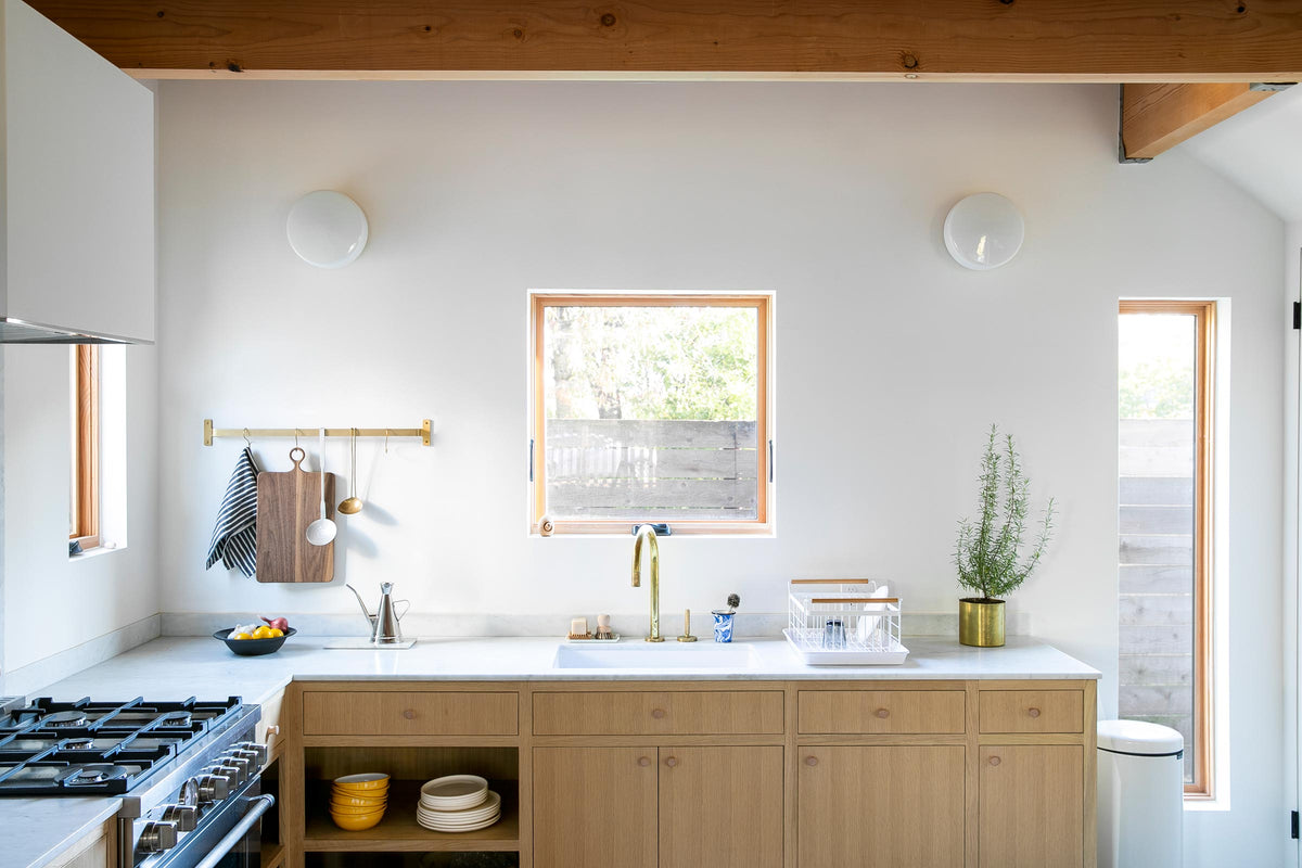 Bright modern kitchen with exposed wooden ceiling beams, white countertop, brass faucet, oak cabinets, small central window