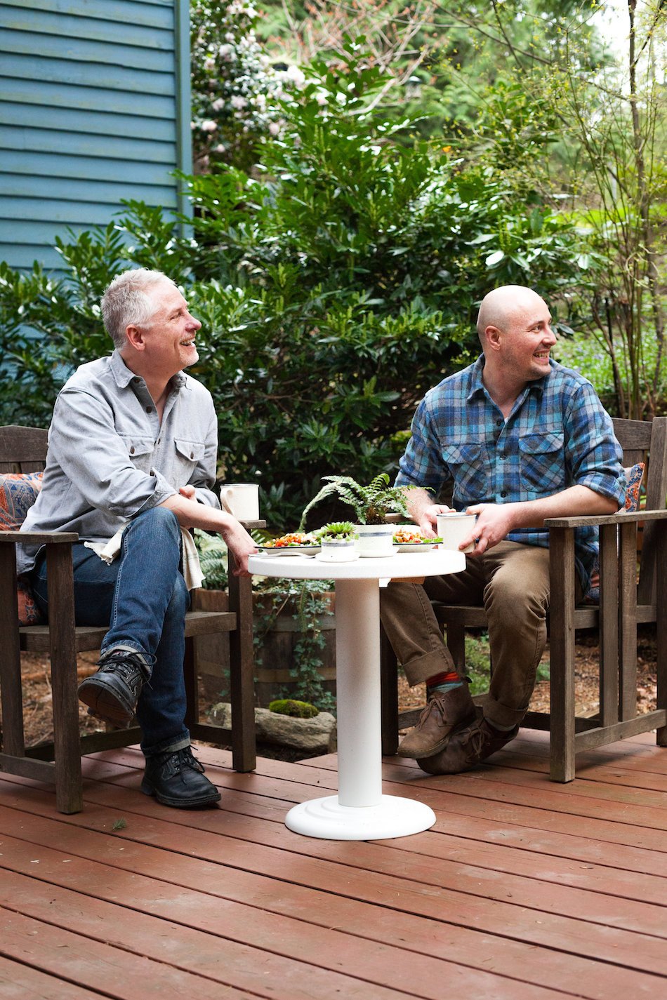 Two men seated on a wooden deck chatting over coffee and snacks at a small white pedestal table in a backyard garden