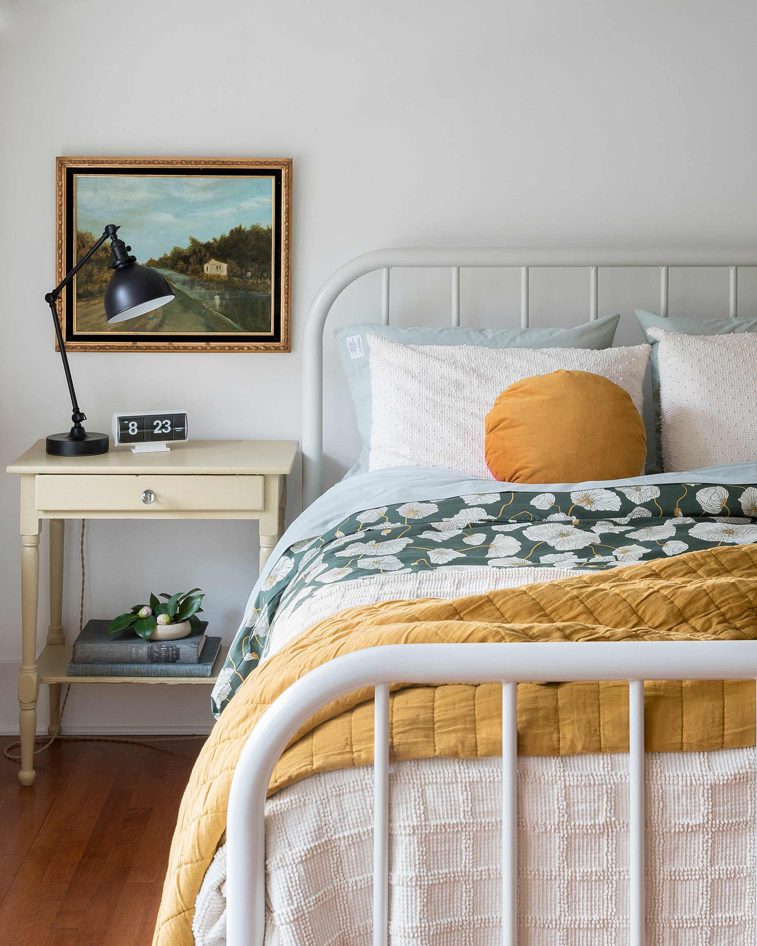 Sunlit bedroom with white metal bed, floral teal duvet, mustard quilt and round pillow, nightstand with black task lamp and framed landscape painting