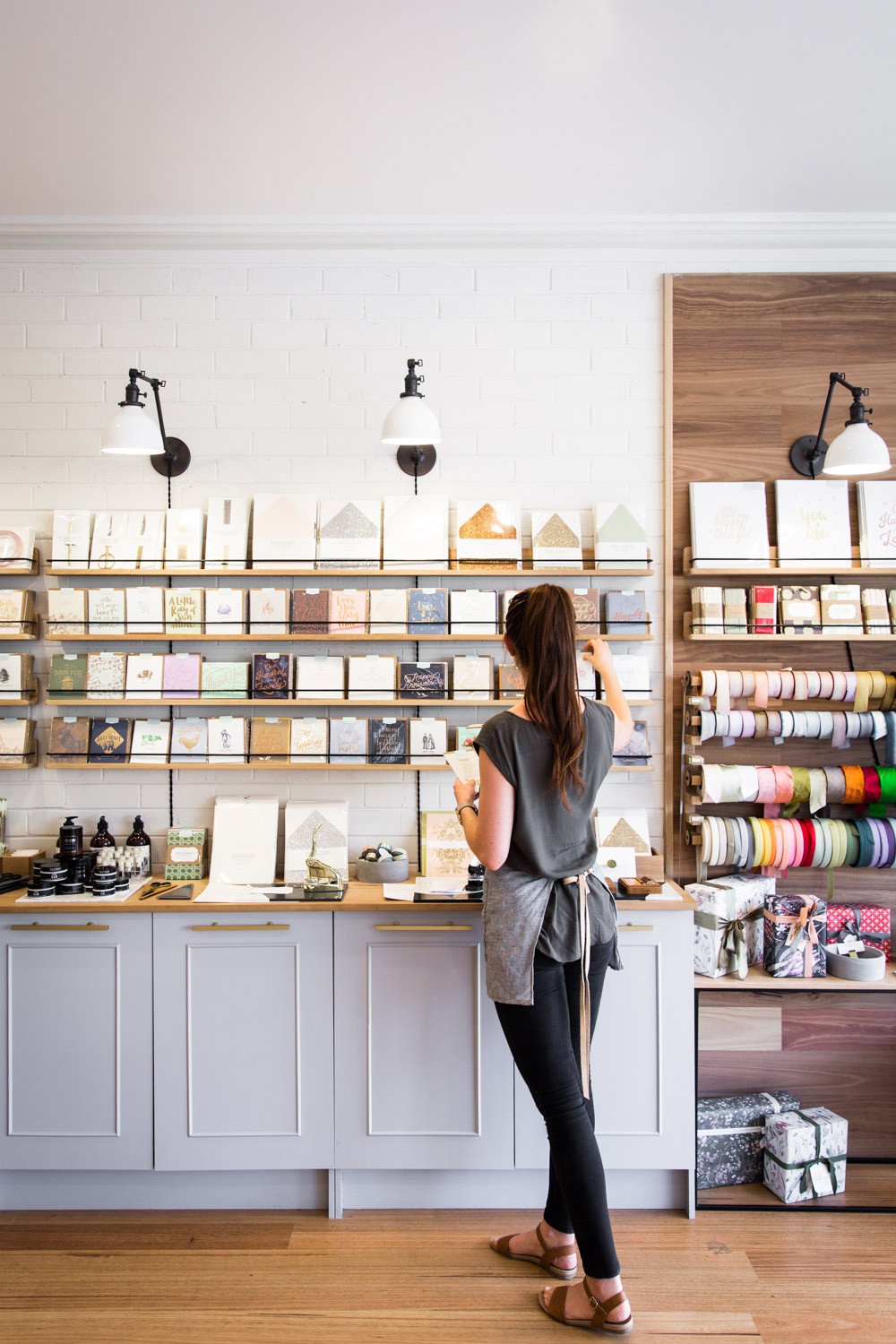 Woman browsing greeting cards on a store display wall with shelves of ribbons and gift wrap in a bright letterpress shop
