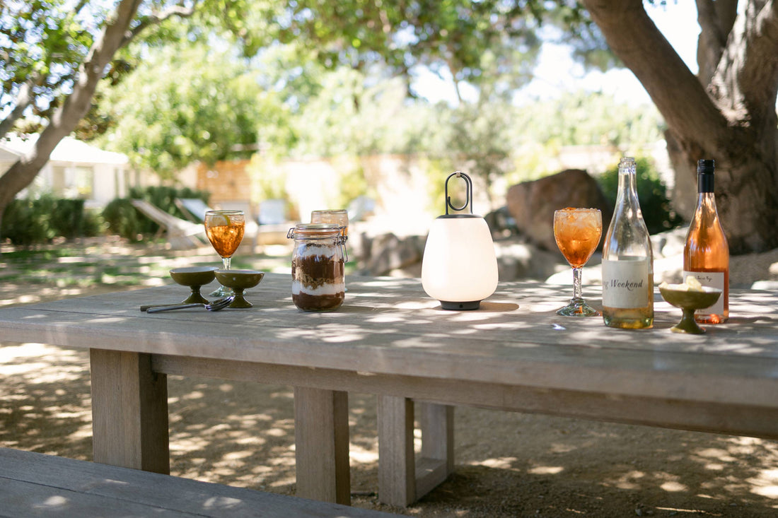 Mason jar of no-bake s’mores icebox cake on a wooden outdoor table beside iced drinks, wine bottles, and a lantern in a shaded backyard
