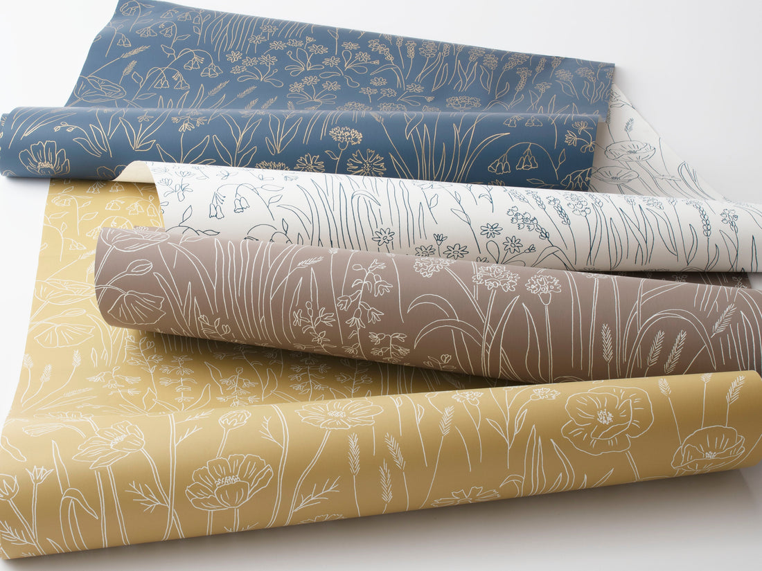 Angled view of stacked wallpaper rolls showcasing a line-drawn botanical pattern in blue, white, taupe, and mustard on a white background