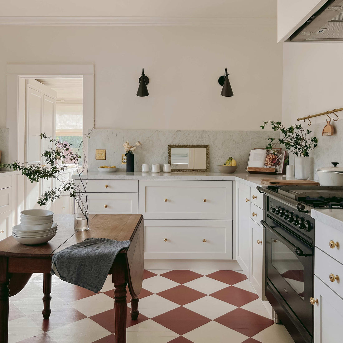 Sunlit kitchen with white shaker cabinets, marble backsplash, wooden island, black cone sconces and red checkerboard floor