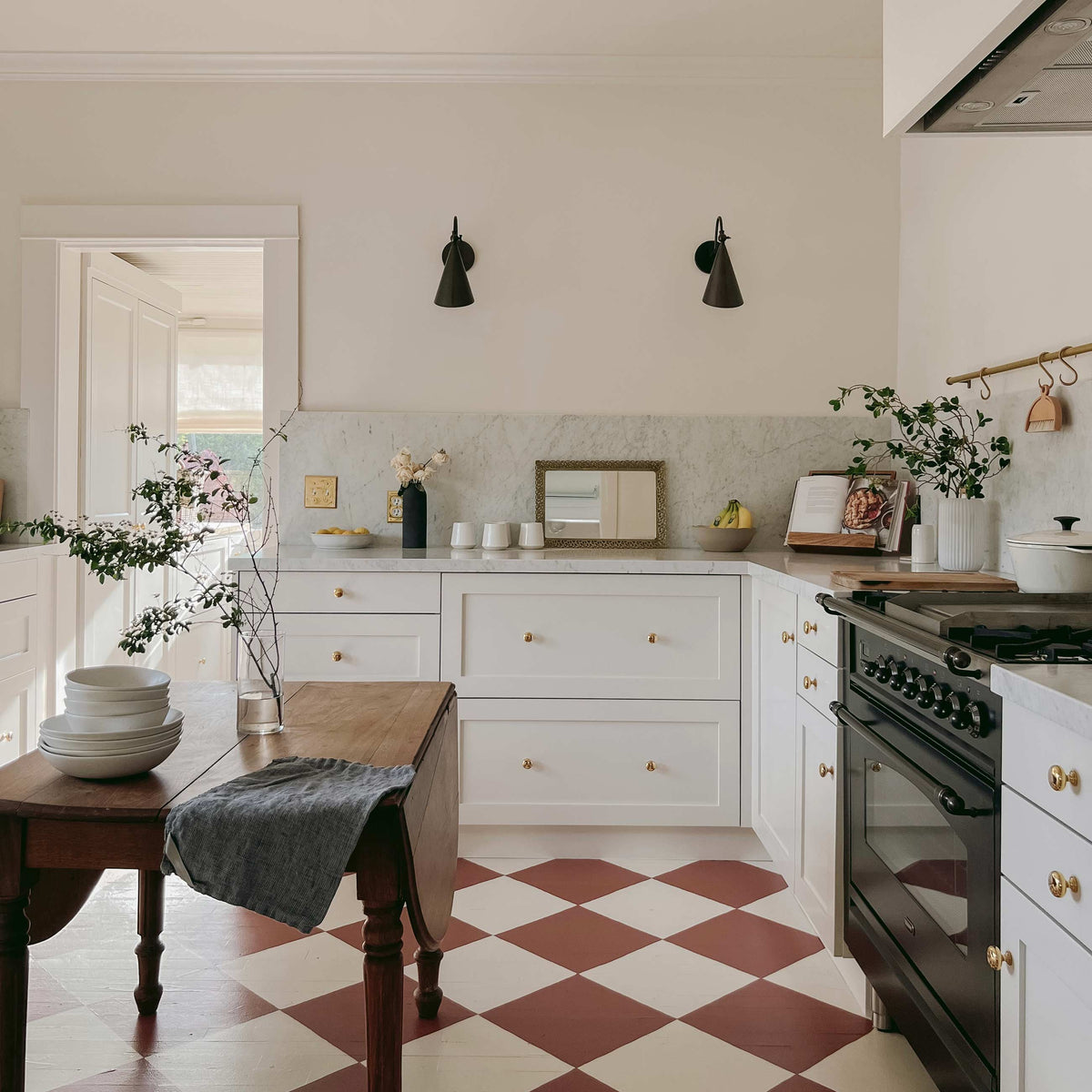 Sunlit kitchen with white shaker cabinets, marble backsplash, wooden island, black cone sconces and red checkerboard floor