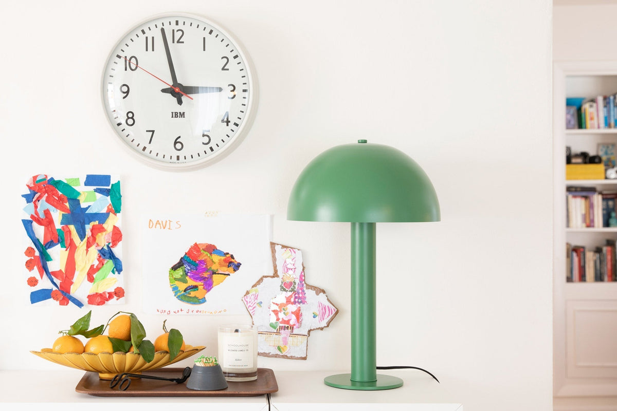 Sidnie lamp in Juniper on a white sideboard beside a bowl of oranges, a candle, and children’s artwork under a wall clock
