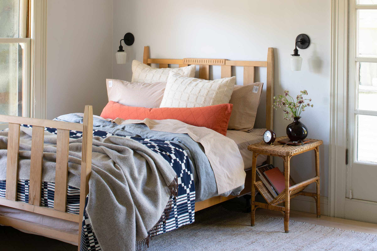 Sunlit bedroom with a wooden bed layered with patterned quilts and pillows, black wall sconces, and a wicker bedside table