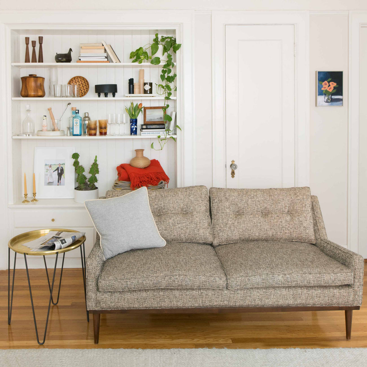 Cozy mid-century style sofa in front of built-in white shelving filled with books, plants, glassware, and decorative objects
