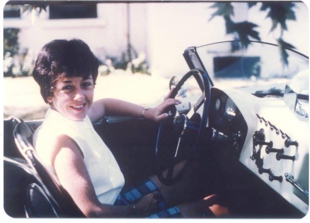Smiling woman seated in a vintage convertible with her hand on the steering wheel, sunlit suburban driveway in the background