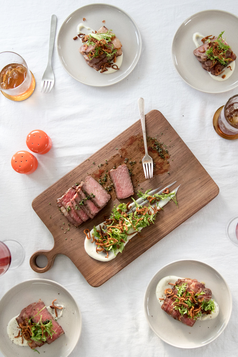 Overhead of sliced roast beef with frisée and fried shallots on a wooden cutting board, plated on a white tablecloth