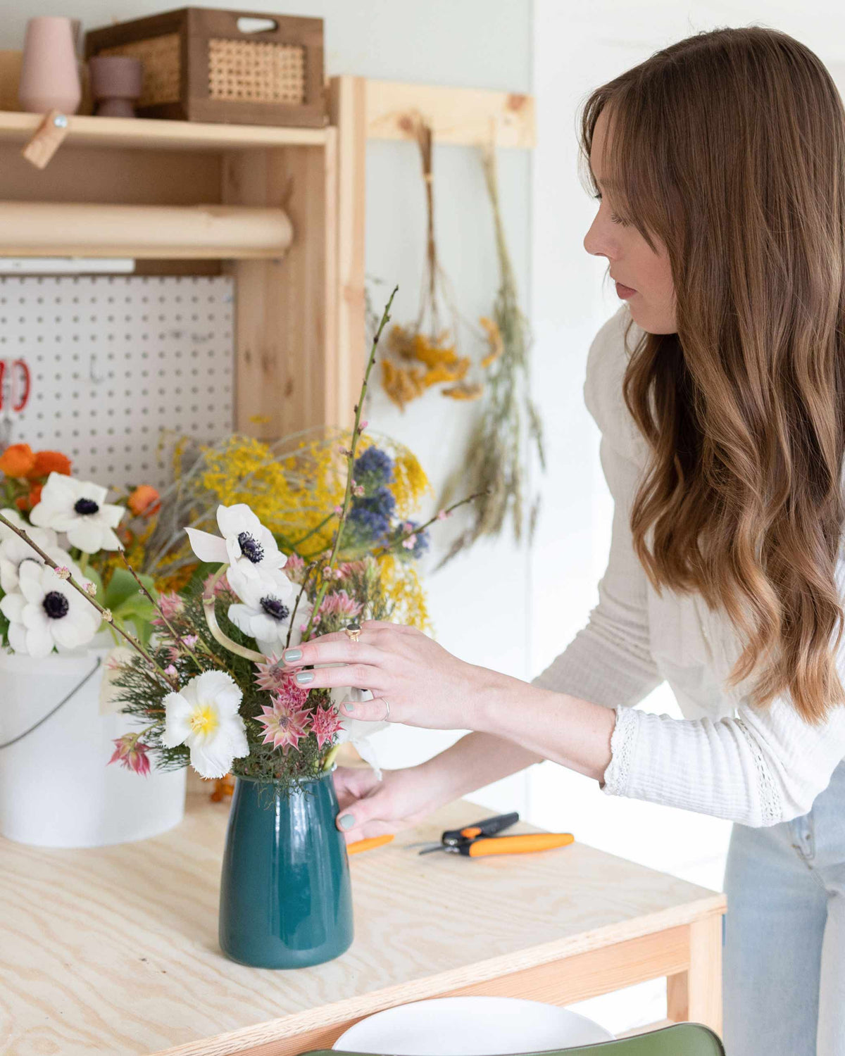 Woman arranging a mixed wildflower bouquet in a teal vase at a bright home workspace