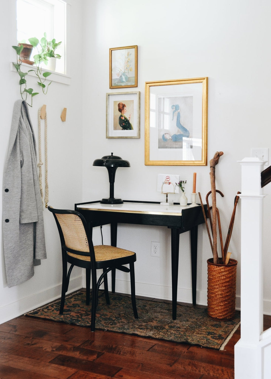 Corner workspace with small black desk and cane chair beneath a gallery wall, coat hooks and woven umbrella stand in a bright white entryway