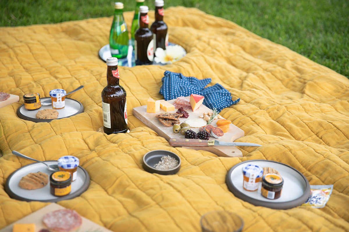 Picnic spread on mustard-yellow quilted blanket with charcuterie board, jars of preserves, crackers, beer bottles, and plates on grass