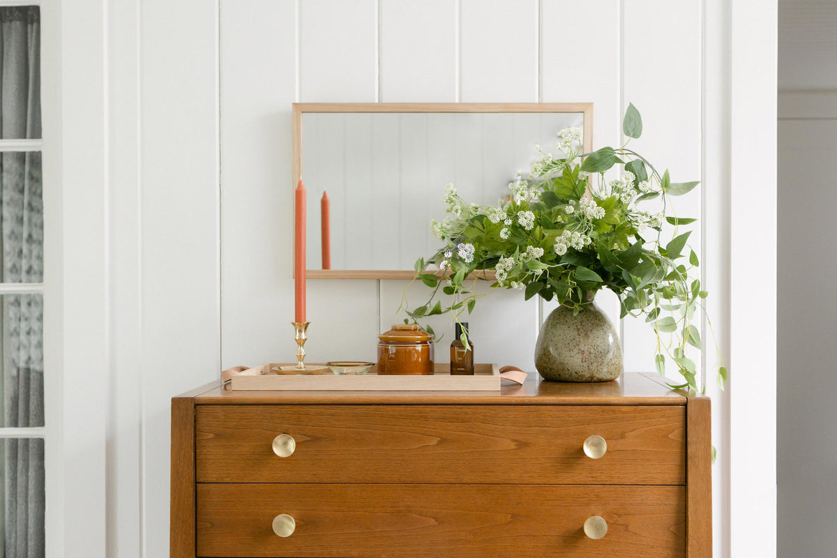 Wood dresser with mirror, coral taper candles on a tray, and a ceramic vase of trailing greenery against white paneled wall