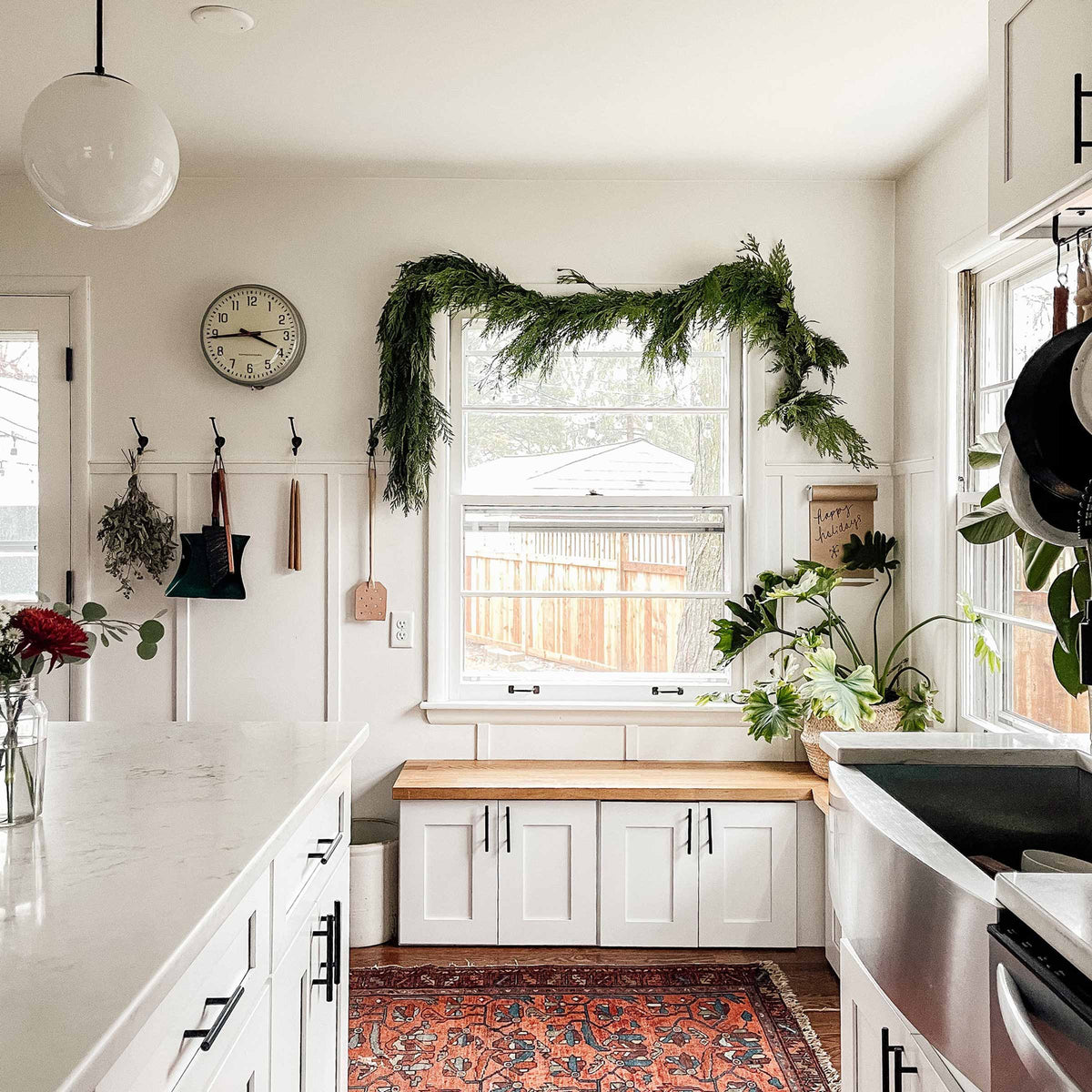 Bright Cape Cod kitchen with cedar garland draped over the window, white shaker cabinets, wood bench, marble island, globe pendant light and potted plants
