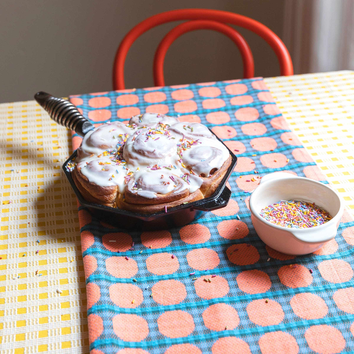Skillet of iced confetti cinnamon rolls sprinkled with rainbow sprinkles on a patterned tablecloth, small bowl of extra sprinkles at right