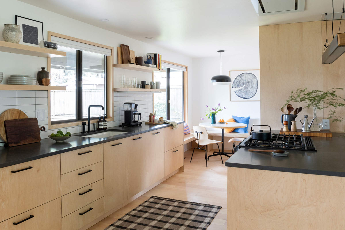 Bright modern kitchen with open shelving, birch cabinets, dark countertops, gas cooktop, and a sunny breakfast nook by the windows
