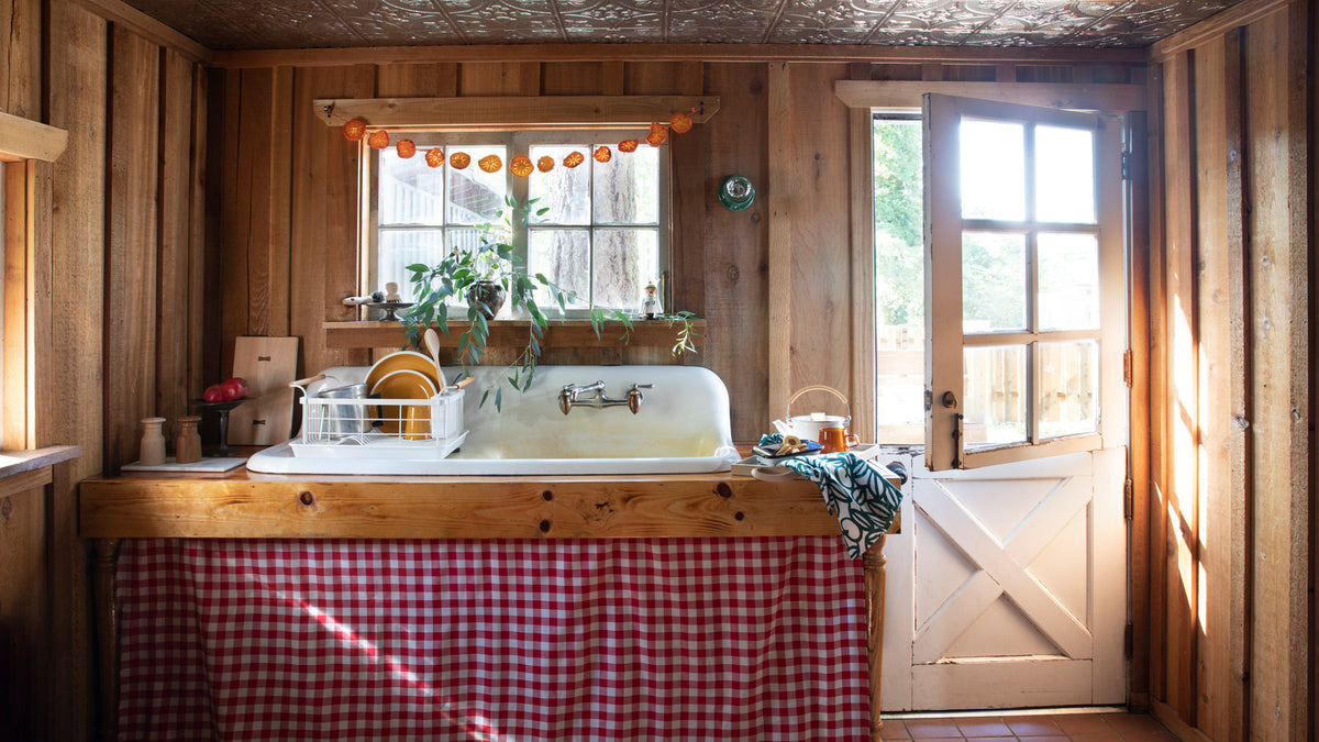 Farmhouse sink with yellow dishes in a wood-paneled cabin kitchen, sunlight coming through a half-open Dutch door