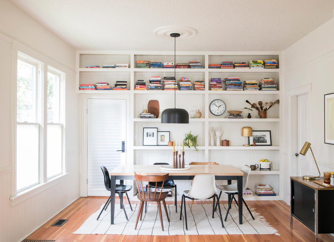 Dining table with mixed chairs and black dome pendant light in a bright white dining room with built-in bookshelves holding stacked books and decorative objects including a small brass table lamp
