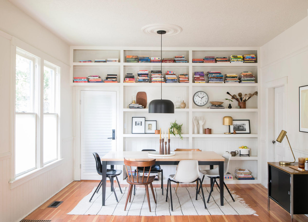 Dining table with mixed chairs and black dome pendant light in a bright white dining room with built-in bookshelves holding stacked books and decorative objects including a small brass table lamp