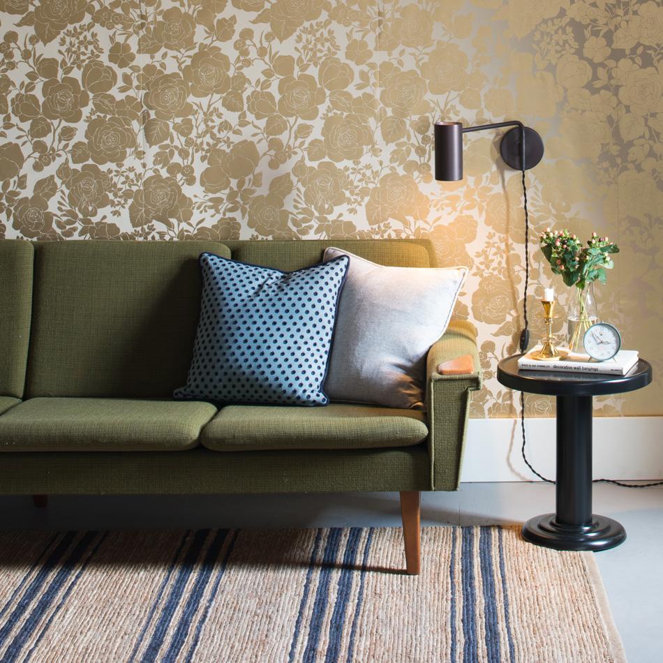 Green mid-century sofa against floral gold wallpaper with wall sconce and black side table holding books a clock and flowers