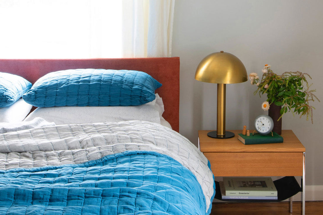 Bed with turquoise quilt and pillows next to a wooden nightstand with a brass dome lamp, small clock, and vase of flowers
