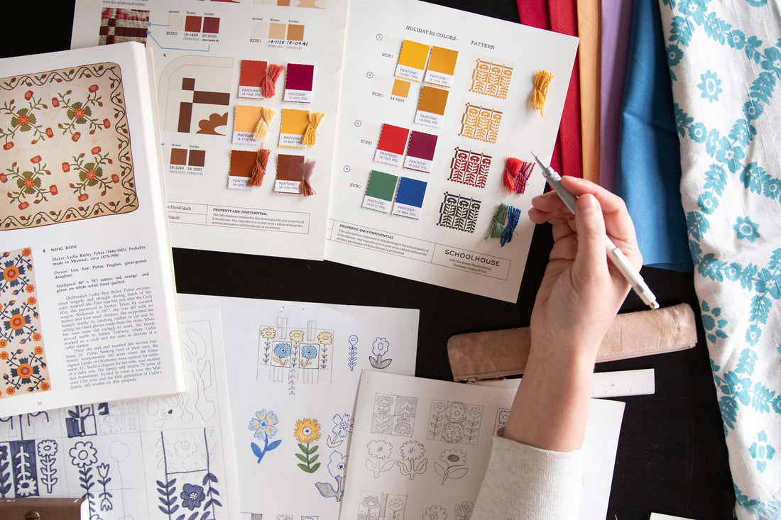 Designer sketching floral quilt motifs at a table with Pantone color swatches, embroidery thread samples, and folded fabric