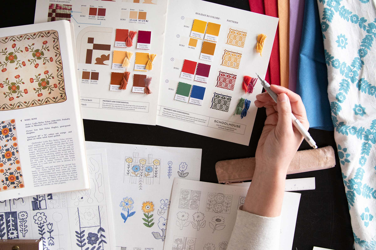 Designer sketching floral quilt motifs at a table with Pantone color swatches, embroidery thread samples, and folded fabric