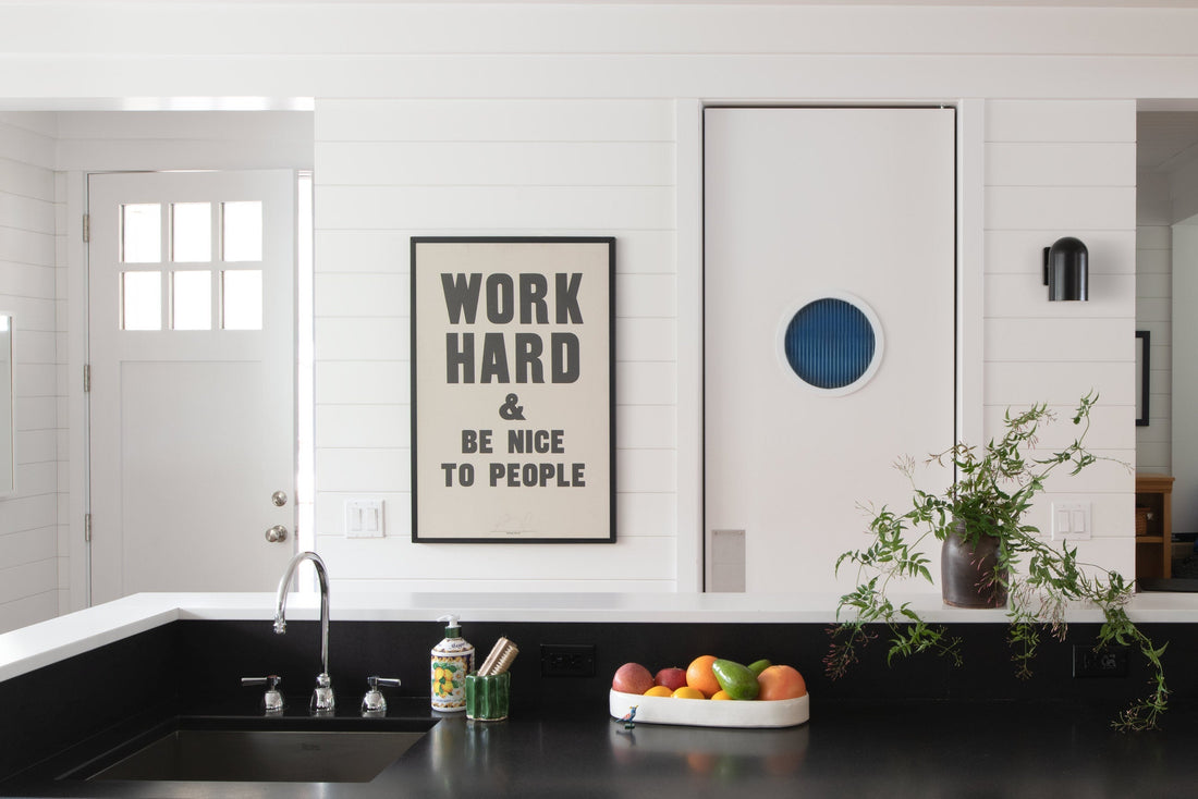 Kitchen counter with black sink and fruit bowl, framed poster reading “WORK HARD & BE NICE TO PEOPLE” on white shiplap wall and white sliding door with blue porthole