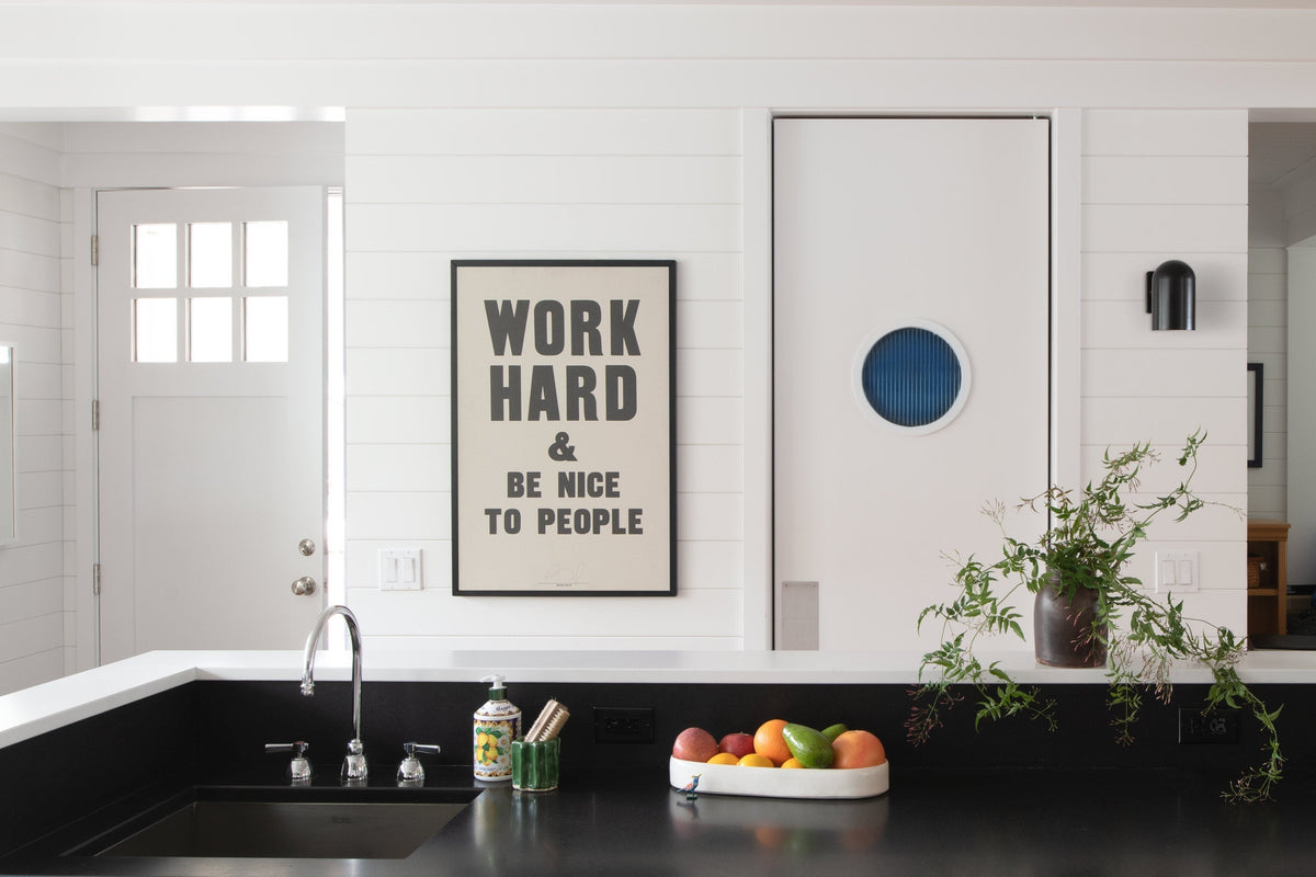 Kitchen counter with black sink and fruit bowl, framed poster reading “WORK HARD & BE NICE TO PEOPLE” on white shiplap wall and white sliding door with blue porthole